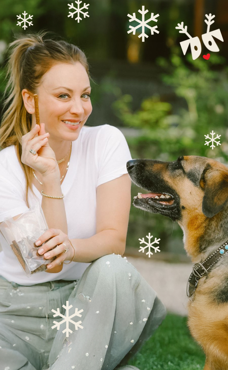 Kaley Cuoco kneeling on grass holds a dog treat toward a large mixed-breed dog; decorative snowflake graphics overlay.