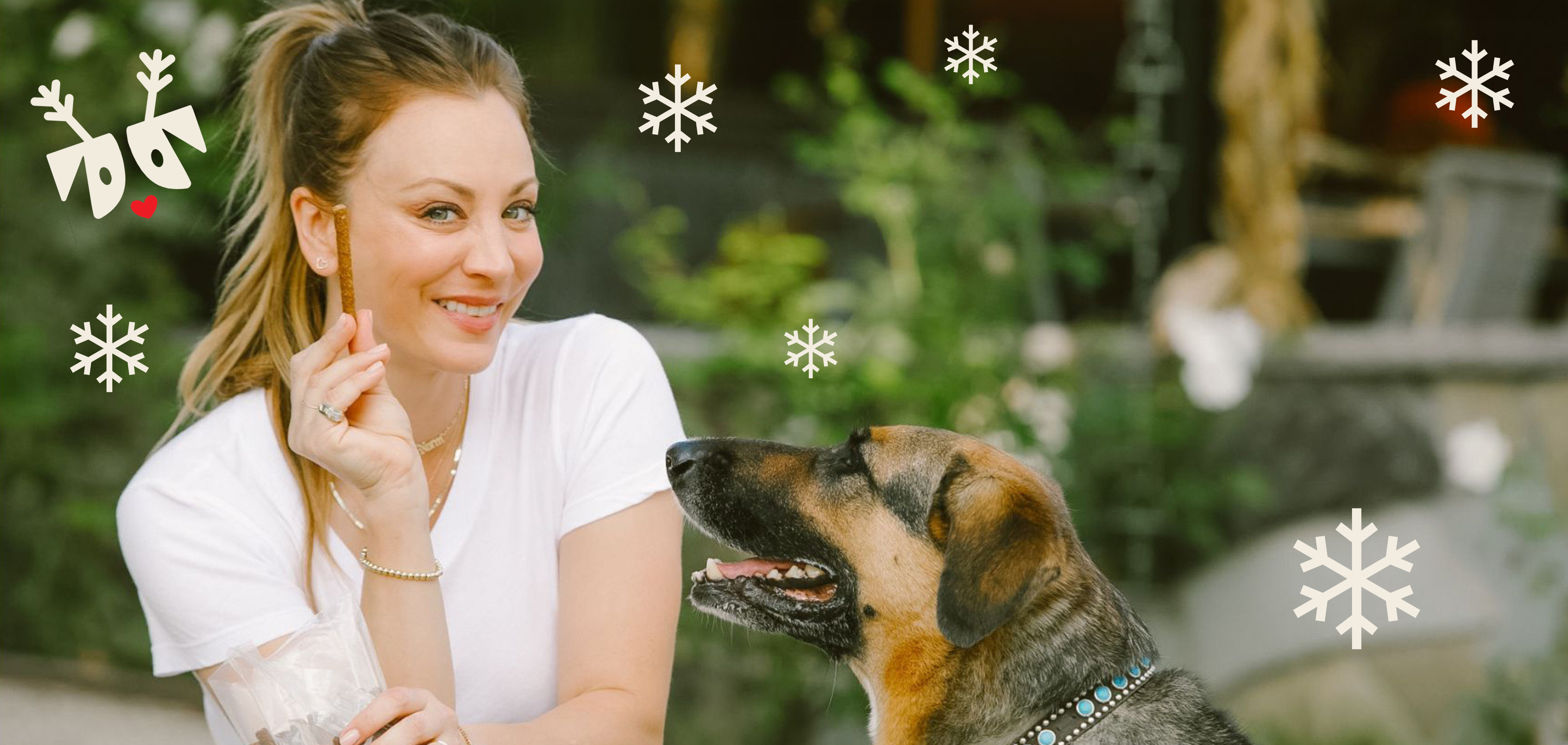 Kaley Cuoco holding a dog treat while a large mixed-breed dog looks up; decorative snowflakes overlay the scene.