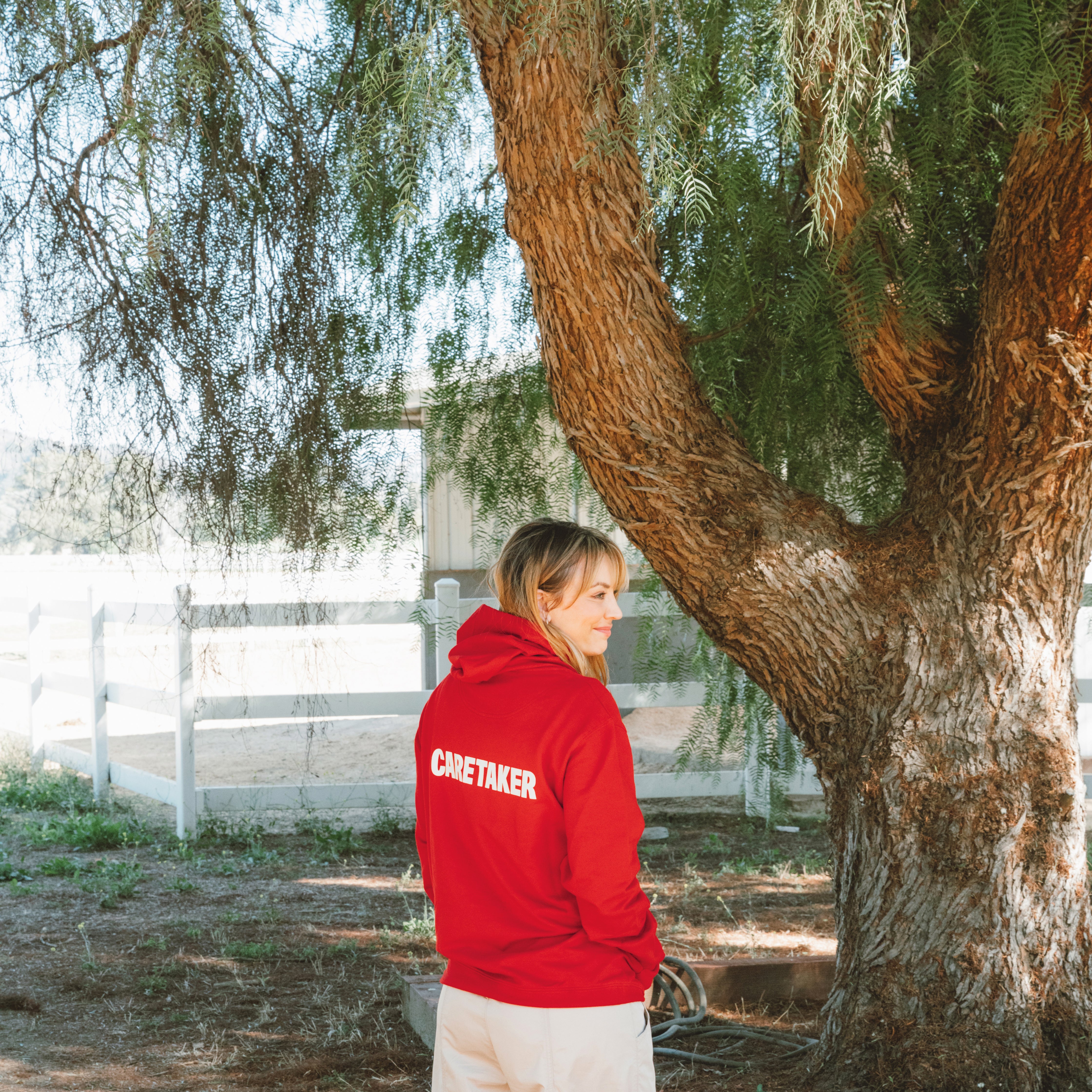 Kaley Cuoco wearing a red hoodie with 'Caretaker Human' text on the back, standing near a tree outdoors.