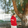 Kaley Cuoco wearing a red hoodie with 'Caretaker Human' text on the back, standing near a tree outdoors.
