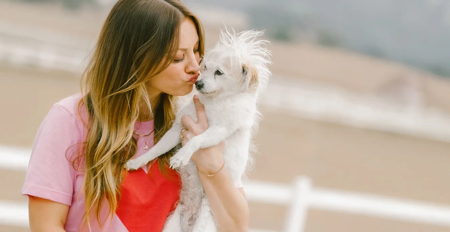 Kaley Cuoco Signature holding and kissing a small white dog outdoors near a white fence, affectionate moment