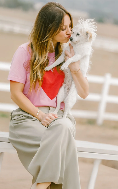 Kaley Cuoco holding and kissing a small white dog on a bench outdoors with a white fence and field in soft focus