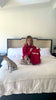 Kaley Cuoco sitting cross-legged on a bed wearing a red hoodie in a bright room