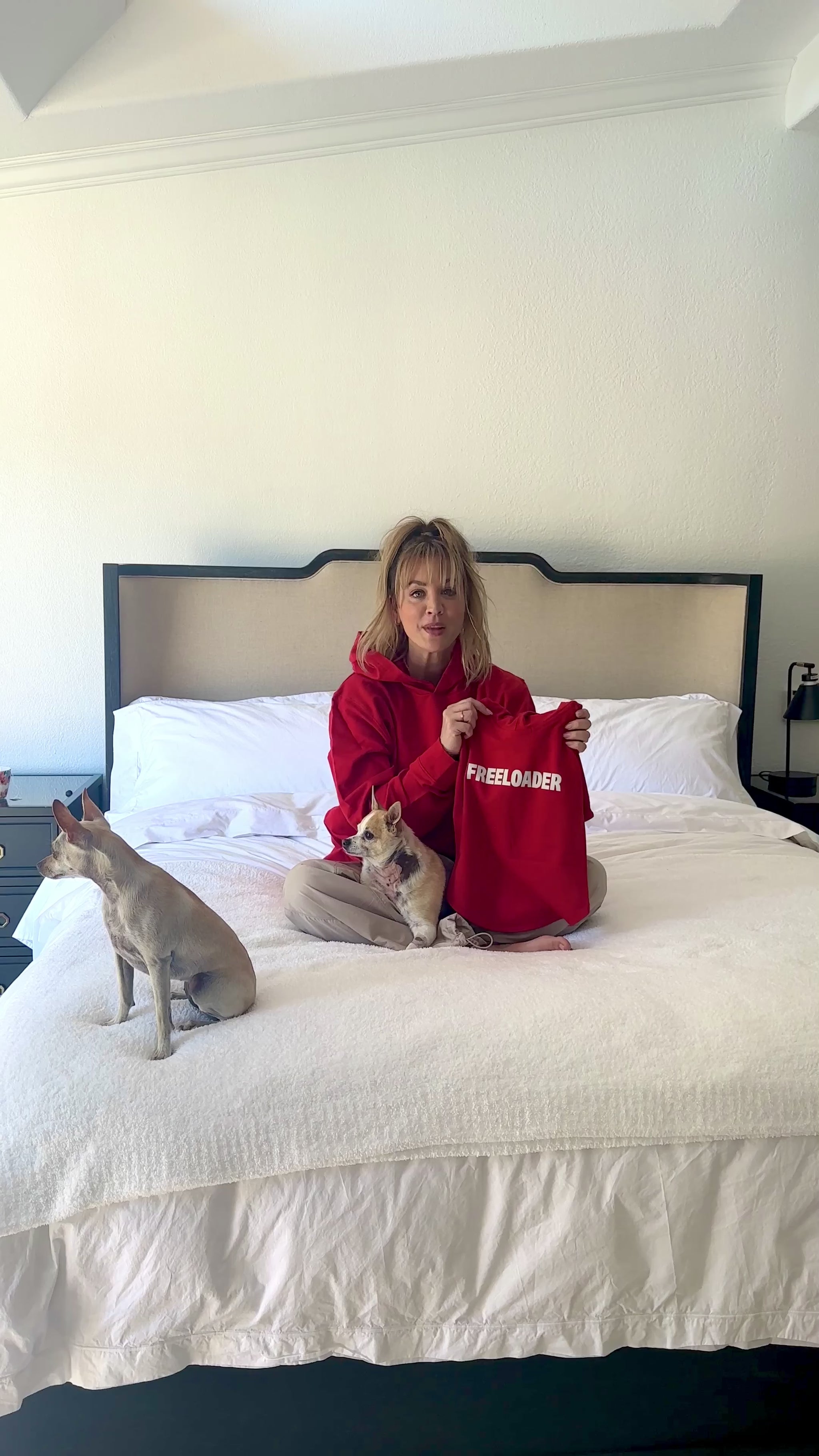 Kaley Cuoco sitting cross-legged on a bed wearing a red hoodie in a bright room