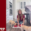 Woman sitting on the floor feeding two dogs from a red Oh-riginal bowl