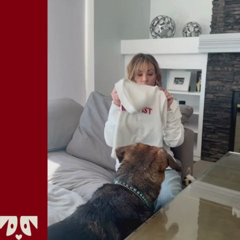 Kaley Cuoco holding a white hoodie with red text 'LOST' facing a dog on a couch