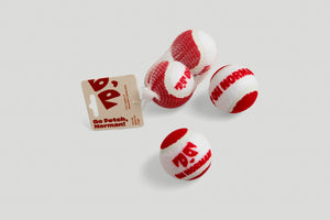 Set of three red-and-white Fetch Me recycled tennis balls in mesh with cardboard tag on light gray background